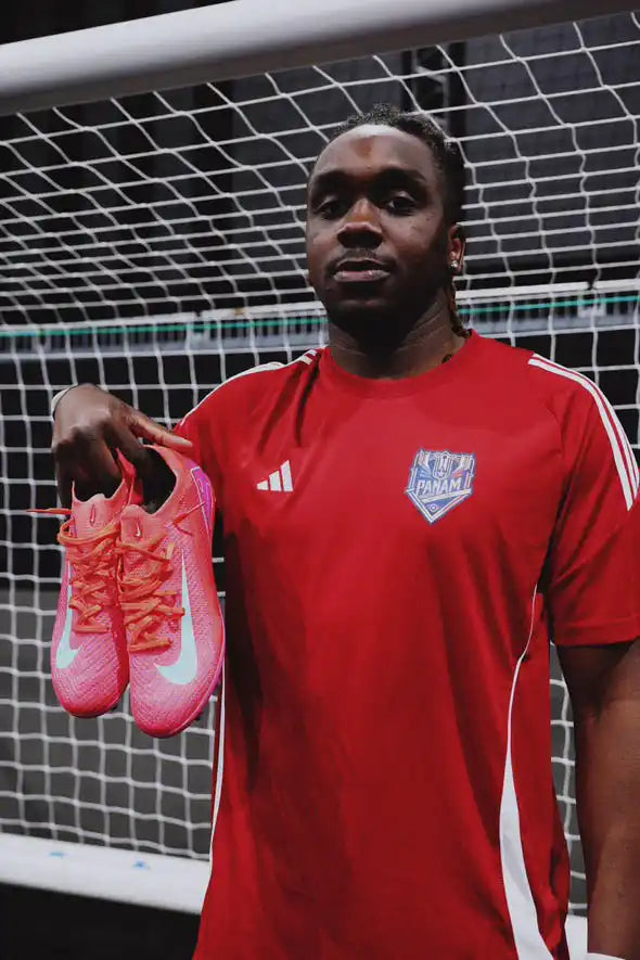 Homme en t-shirt de sport rouge tenant une paire de chaussures de soccer roses.