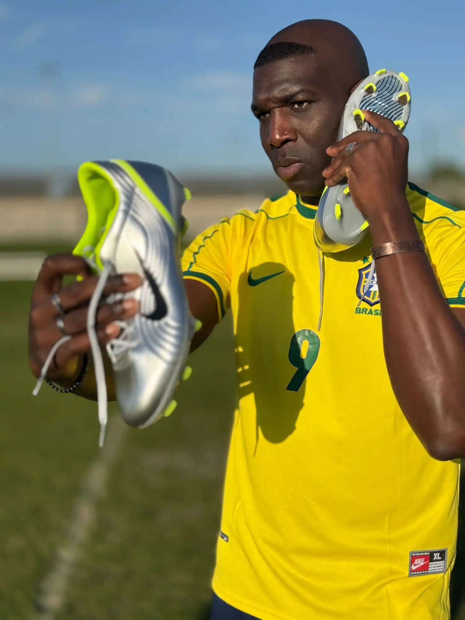 Un homme à la peau sombre portant un maillot jaune tenant deux chaussures de football argentées et vert lime.